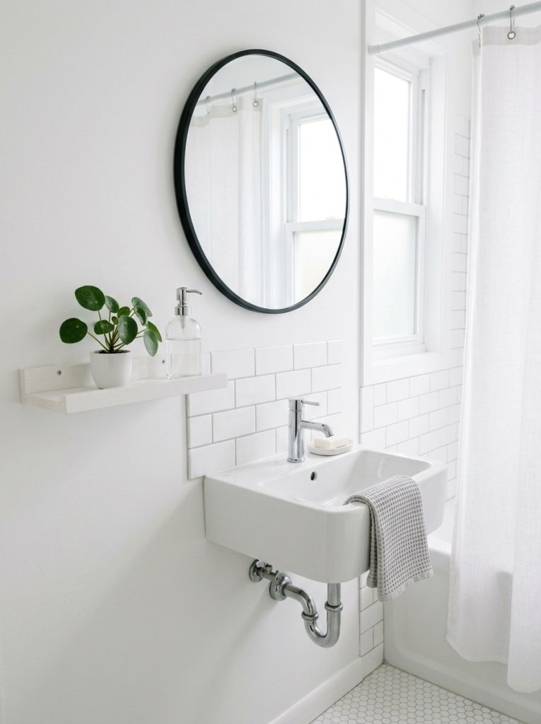 Clean small bathroom aesthetic with white walls tiles chrome faucet narrow shelf plant soap dispenser and round mirror