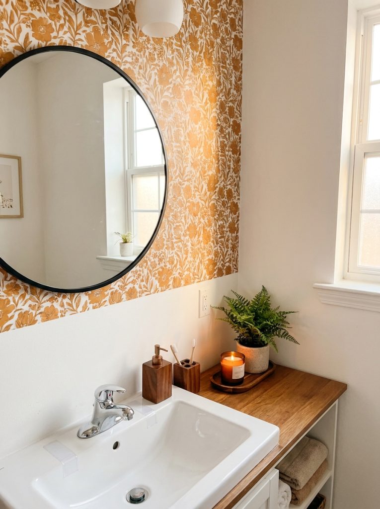 Cozy apartment bathroom aesthetic with white walls black mirror brown wood accessories warm wallpaper candle and plant